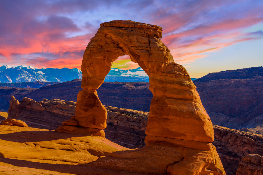 A natural sandstone arch with a colorful sunset sky and snowy mountains in the background.