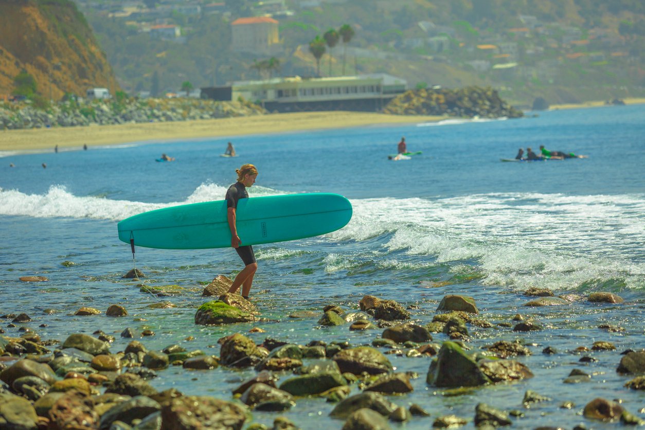 Rocky coastline, surfer with turquoise board foreground, distant cliffs/buildings, midday light, editorial travel photo, people.
