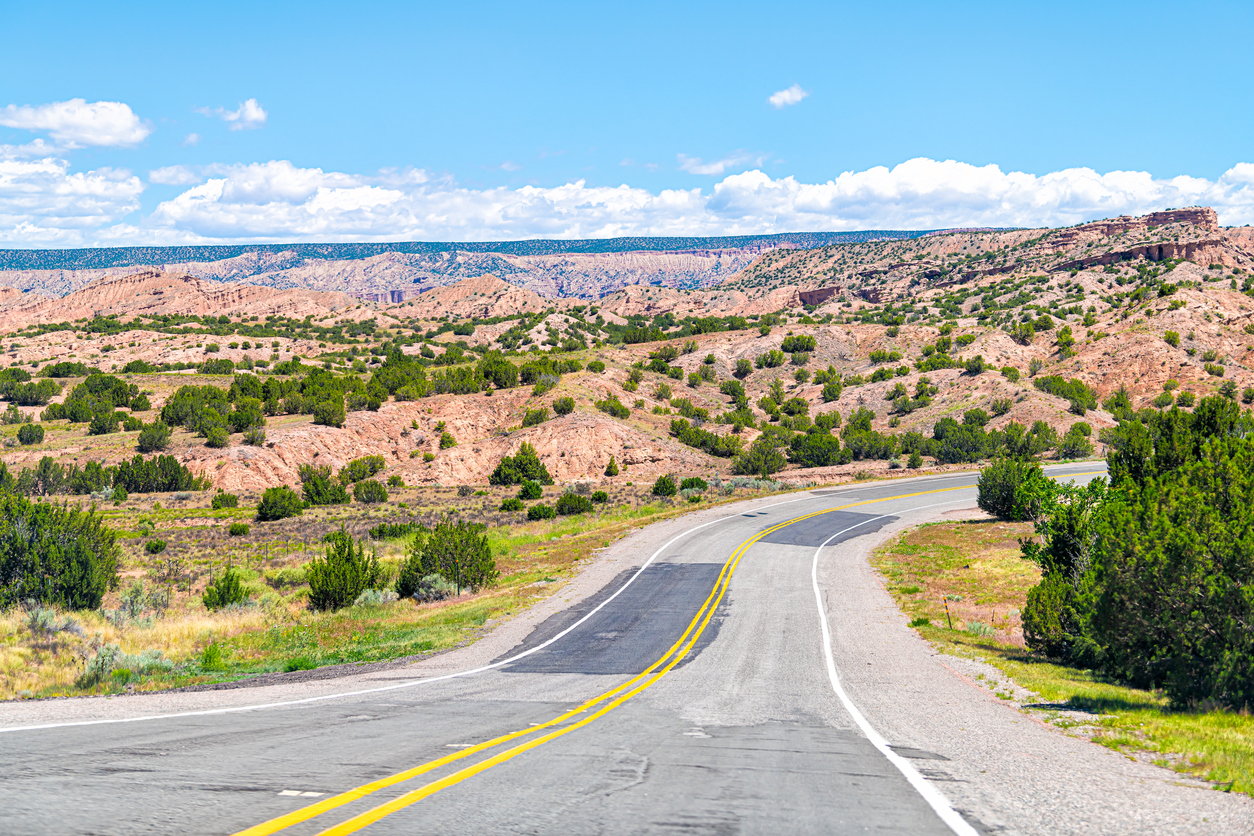 Curved road through desert landscape with shrubs, hills, and a partly cloudy sky.