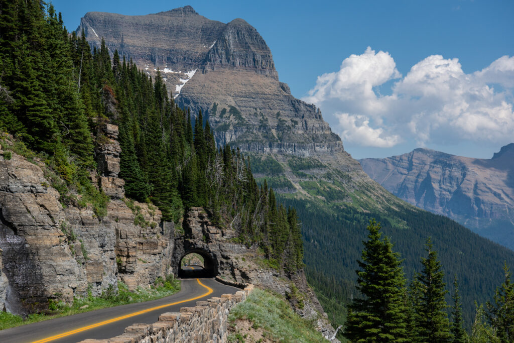 Mountain pass, curved tunnel, pine forest foreground, daytime with scattered clouds, editorial travel photo, no people.
