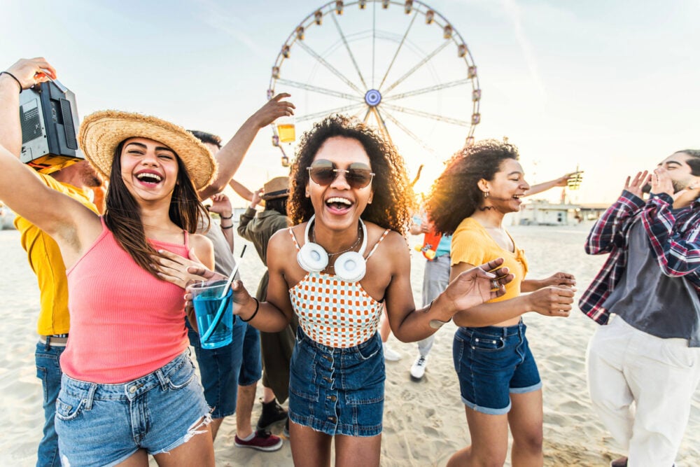 Group of people smiling and dancing on a sandy beach with a Ferris wheel in the background.