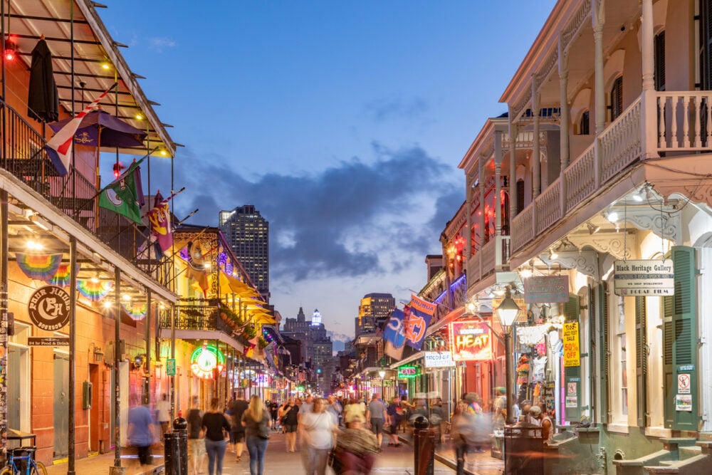 Bustling street scene at dusk, colorful signs and flags, people walking, buildings with balconies and lights.