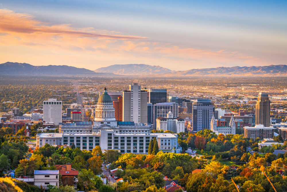 Skyline of a city with a prominent domed building, surrounded by trees, against a mountain backdrop at sunset.