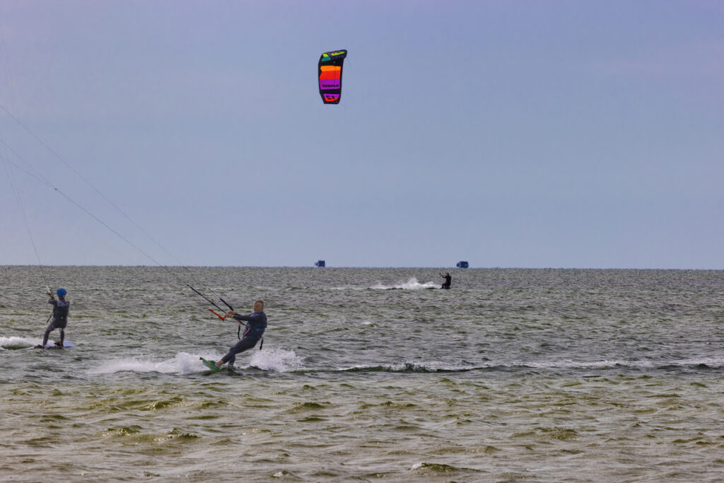 Beach, two kite surfers and colorful kite, wide view with horizon line, midday clear sky, editorial travel photo, people present.