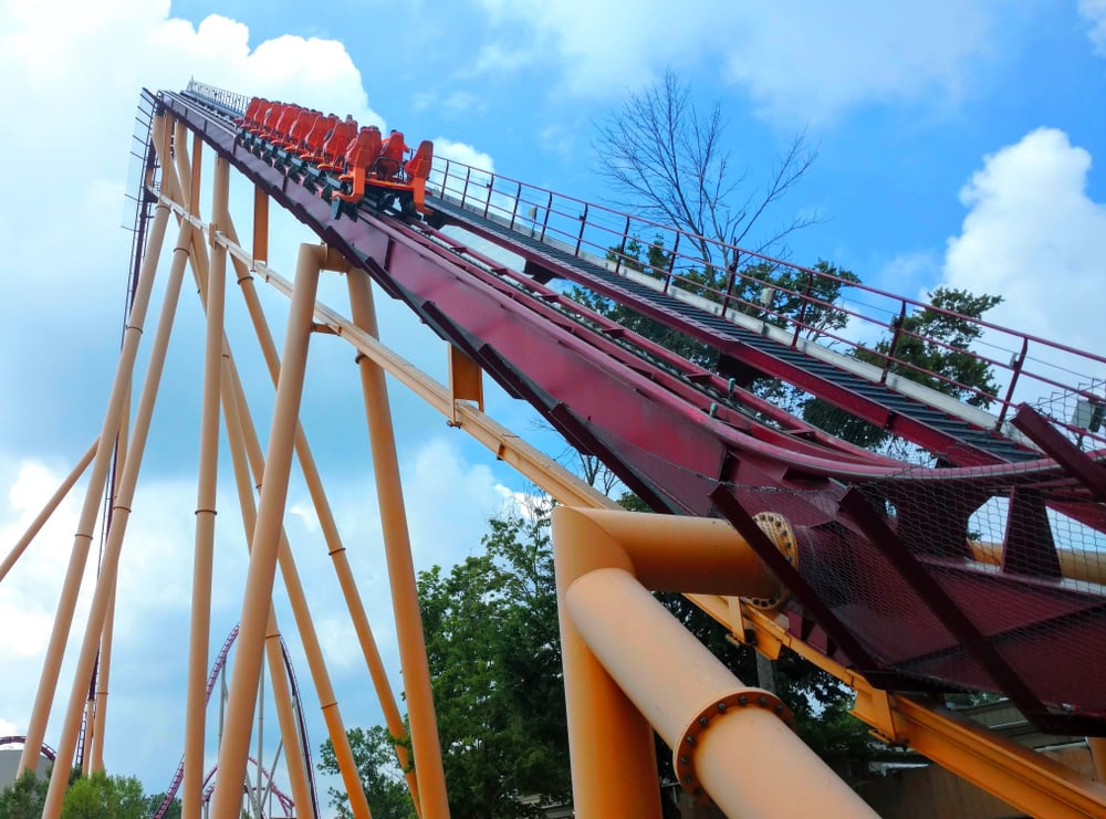 Amusement park, red and yellow coaster climbing, vertical track framed by trees, midday sun, editorial travel photo, with riders.