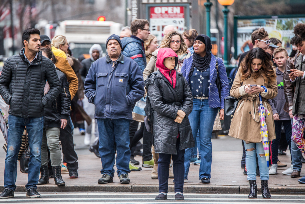 A diverse group of people standing on a city sidewalk near a crosswalk, some looking down at phones.