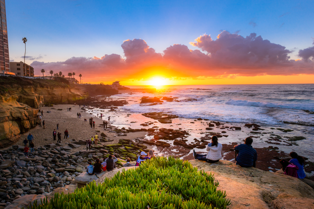 People sitting on rocks and beach watching sunset over the ocean, with clouds and waves.