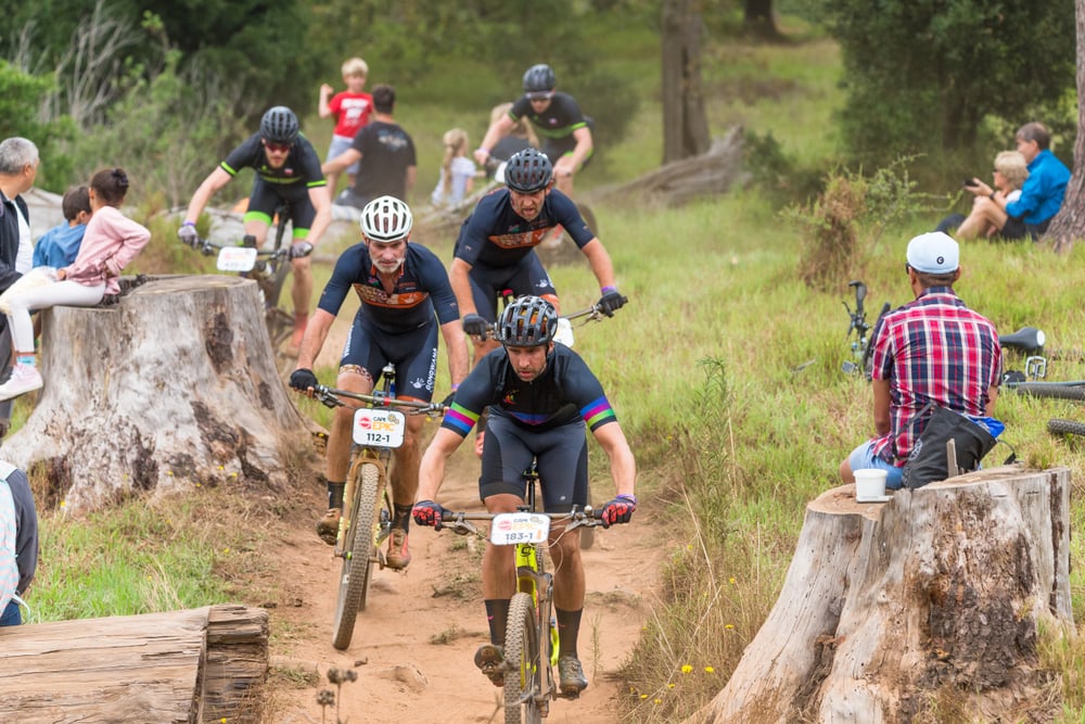 Cyclists riding on a dirt trail, spectators sitting and watching near large tree stumps.