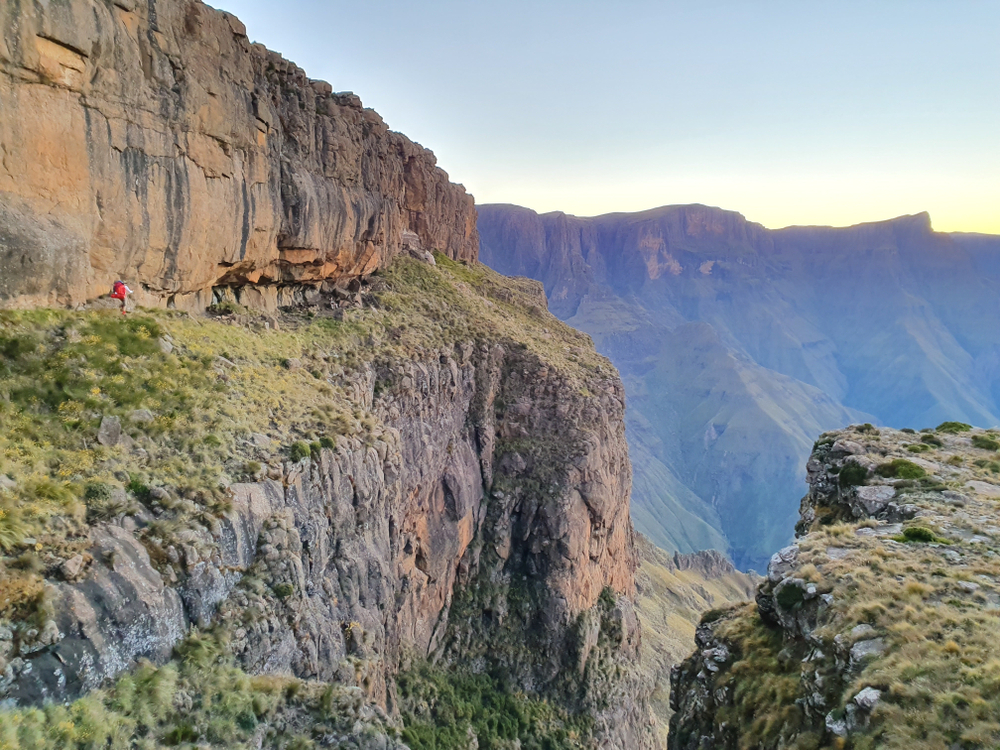 A person in red walks along a rocky cliff edge with a mountainous backdrop at sunrise or sunset.