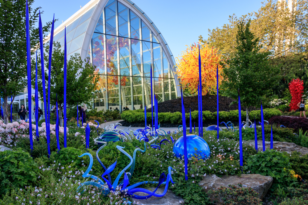 Garden, tall blue glass sculptures, lush plants fronting glass conservatory, daytime sun, editorial travel photo, no people.