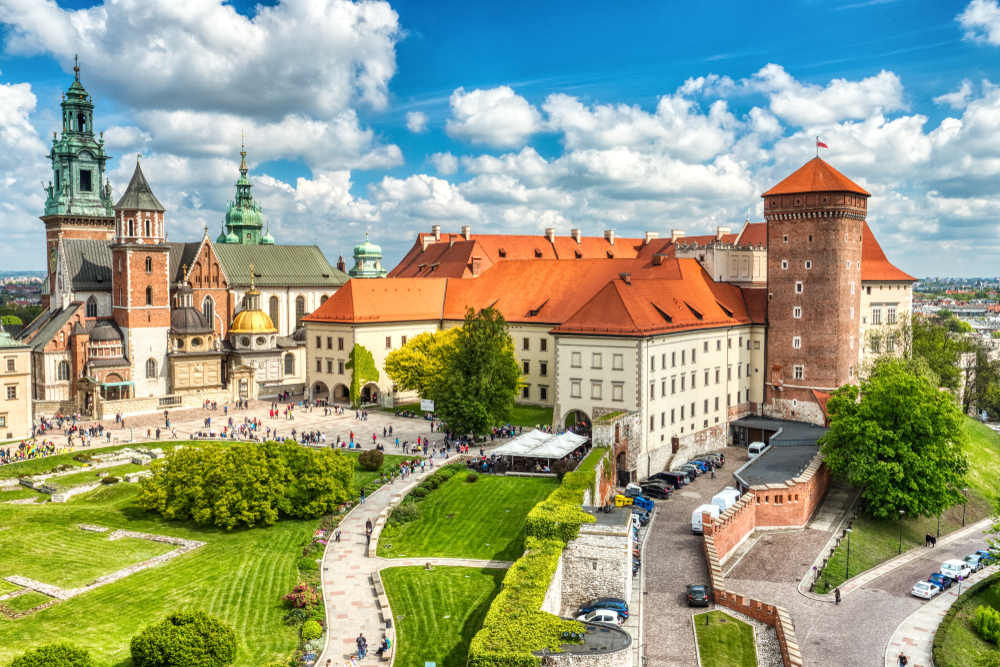 Wawel Castle complex in Krakow, with towers, green domes, red rooftops, and surrounding gardens under a cloudy sky.