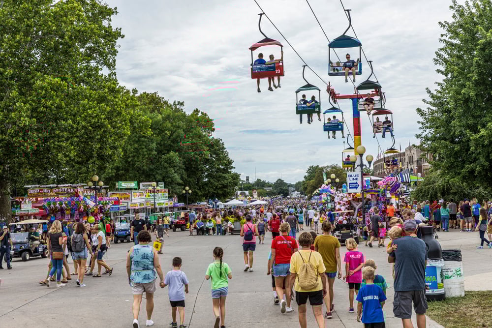 Fairground, chairlift overhead, crowded path flanked by booths, overcast daylight, editorial travel photo, people present.