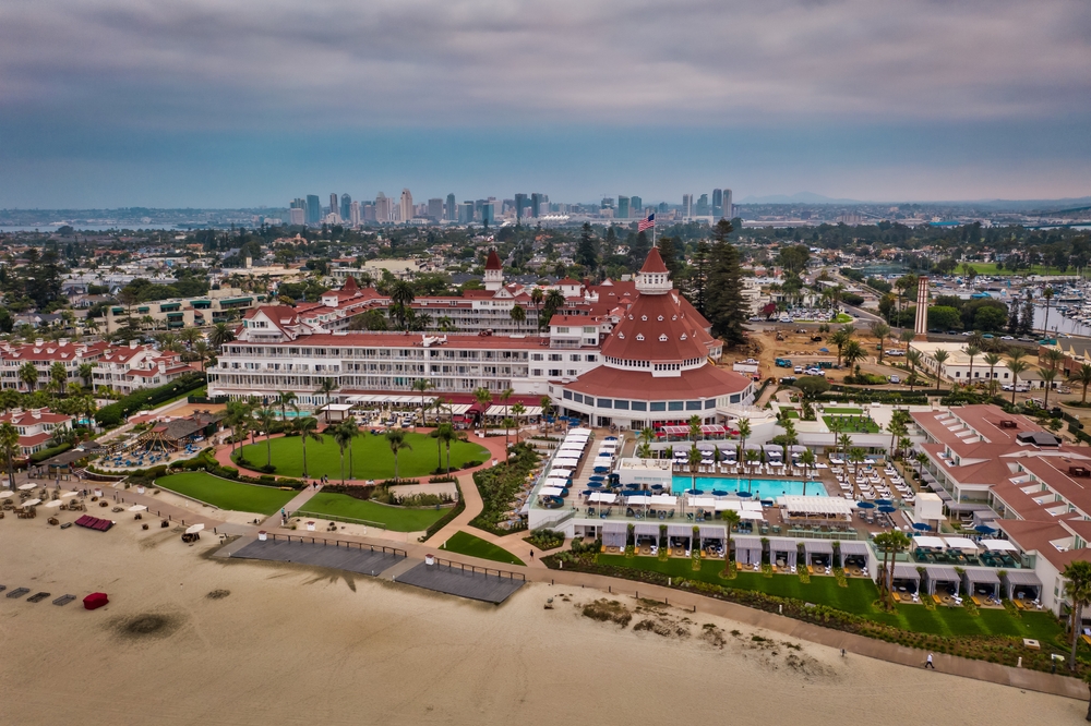 Aerial view of a large coastal hotel with red rooftops, surrounded by greenery and a sandy beach.