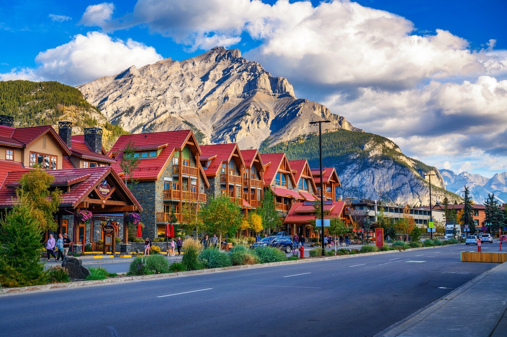A street lined with colorful buildings, trees, and mountains in the background under a partly cloudy sky.