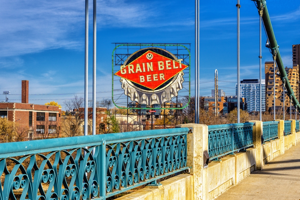 Minneapolis riverfront, iconic Grain Belt Beer sign, viewed from ornate blue bridge, bright midday sun, editorial travel photo, no people.