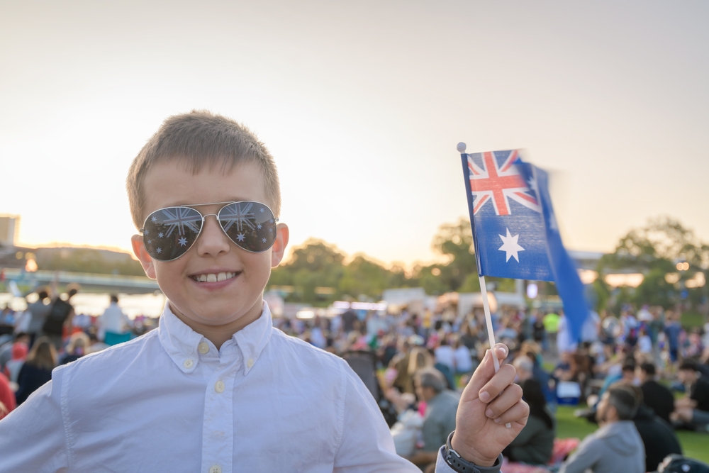 Grassy outdoor festival, boy with sunglasses and Australian flag foreground, sunset glow, editorial travel photo, people present.