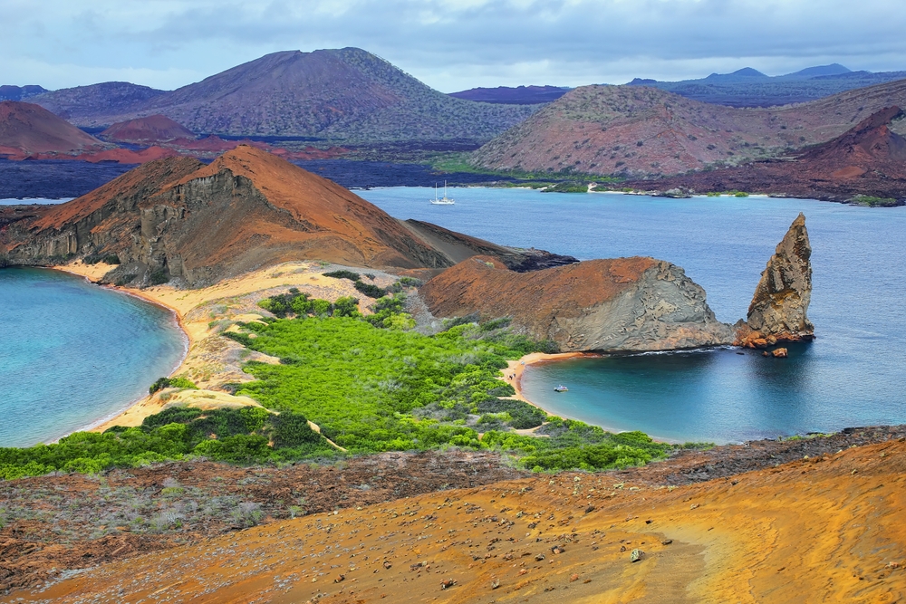 Coastal landscape with volcanic mountains, turquoise water, a rock formation, sand, and green vegetation under cloudy sky.