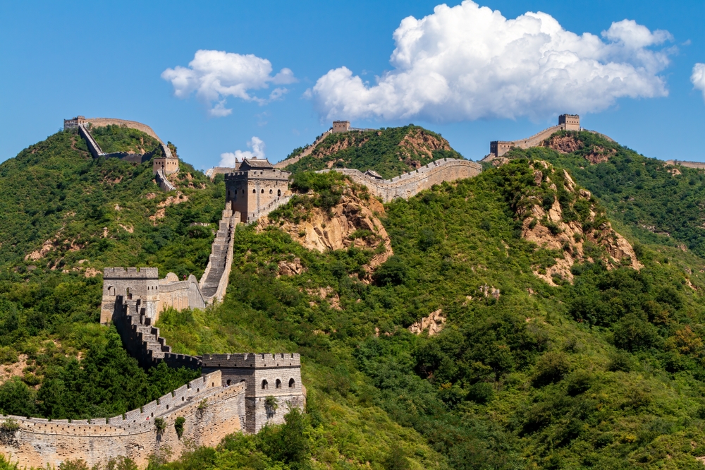 The Great Wall of China stretches across green hills under a blue sky with some clouds.