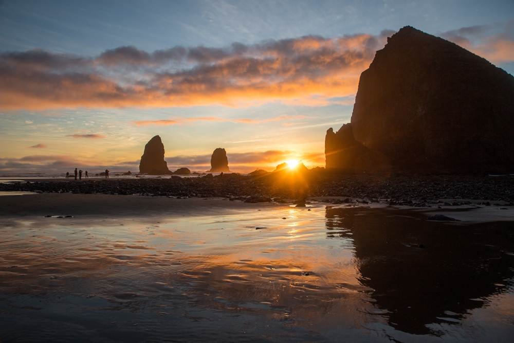 Sunset view with silhouetted rock formations and people on a beach, reflected in wet sand.
