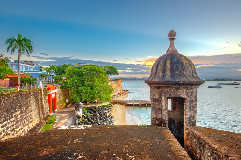 Historic fort walls with a sentry box overlooking a bay at sunset, with palm trees and buildings.
