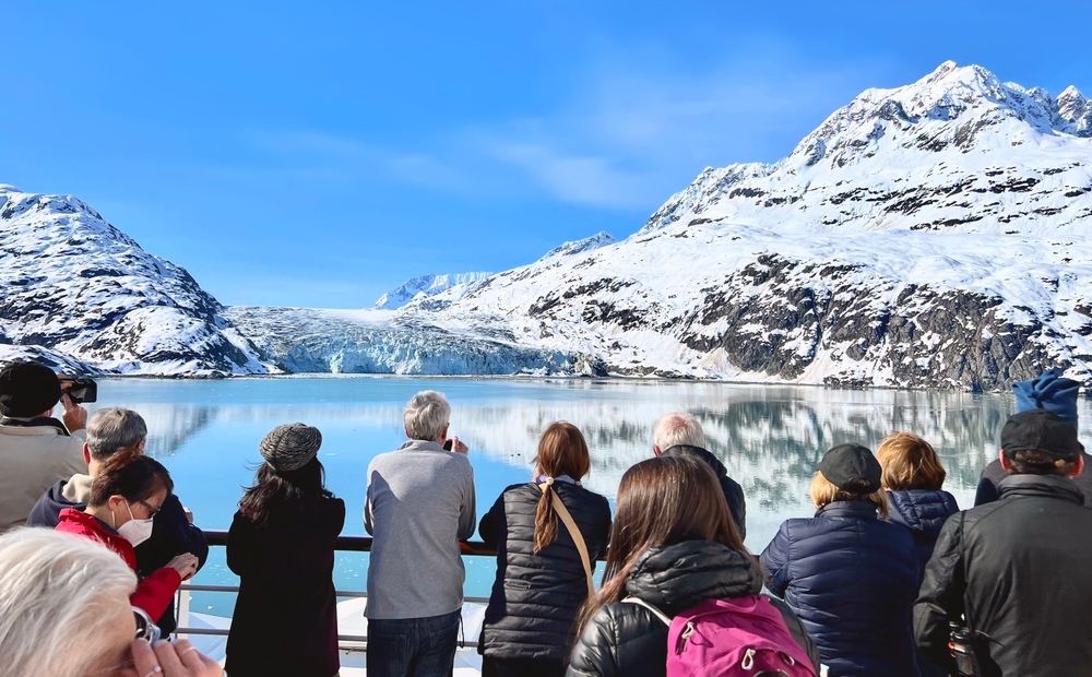 People on a boat observing snowy mountains and a glacier reflected in a calm body of water.