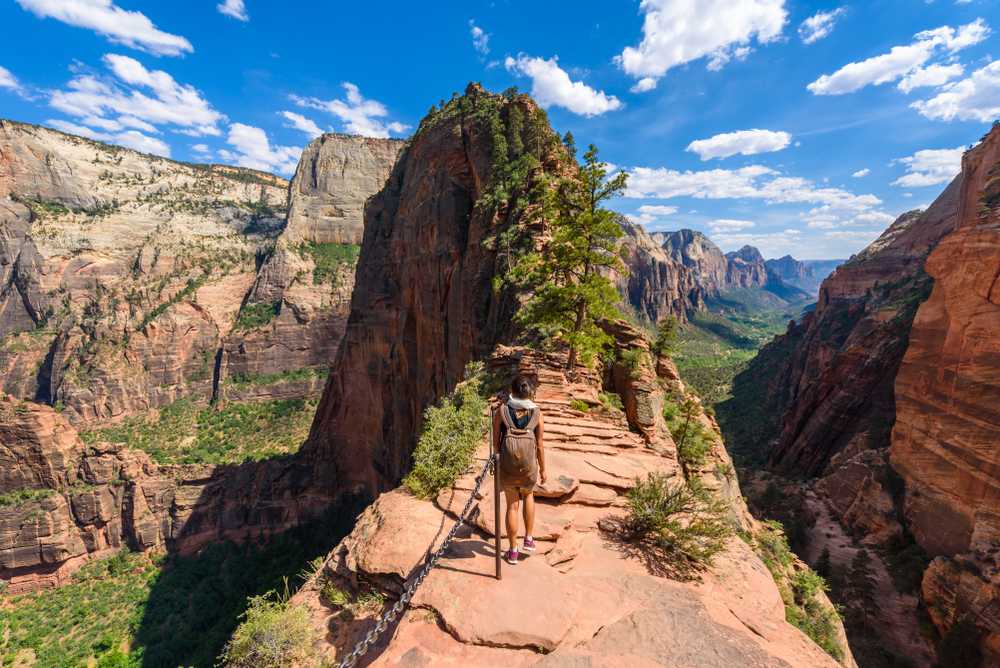 Hiker stands on narrow cliff path surrounded by canyon and mountains under blue sky with scattered clouds.