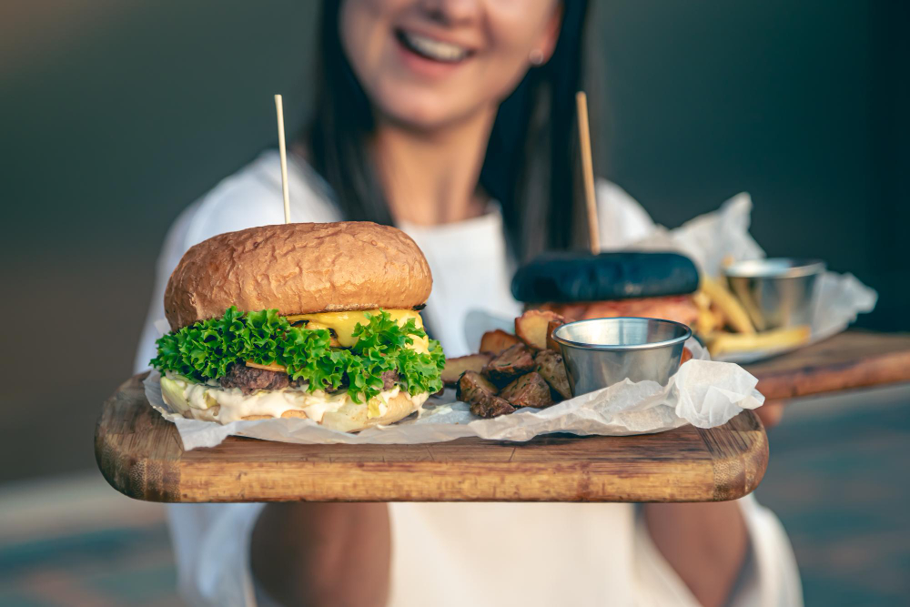 Casual eatery, burgers and sides on boards foreground, smiling woman blurred background, bright daylight, editorial travel photo, people.
