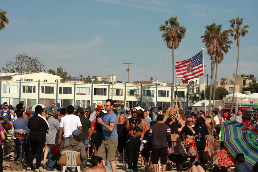 Crowd gathered on a sunny beach, with an American flag and palm trees in the background.