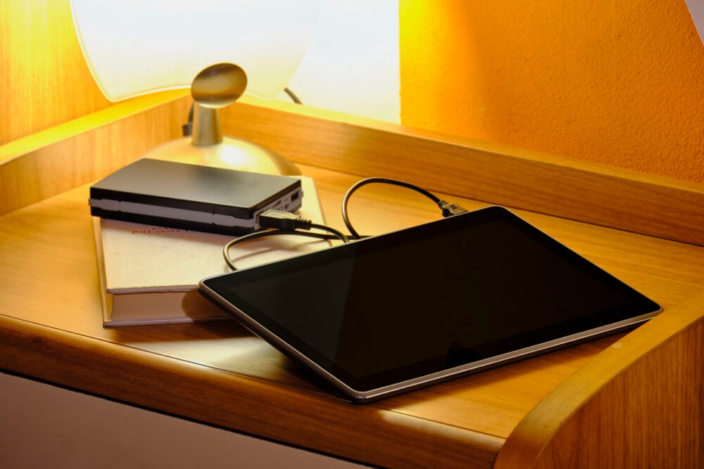Wooden nightstand, tablet with hard drive on closed book, side-lit lamp, evening ambient light, documentary style, no people.