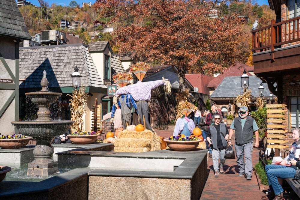 Outdoor plaza, autumnal decorations with scarecrow, pumpkins by fountain, golden daylight, editorial travel photo, people walking.