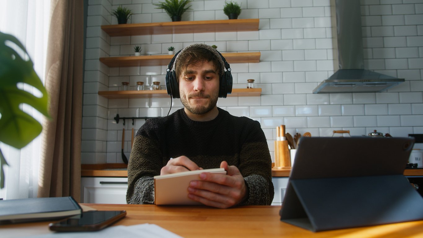 Kitchen table, man with headphones writing, tech devices and papers scattered, morning light, documentary style, one person.