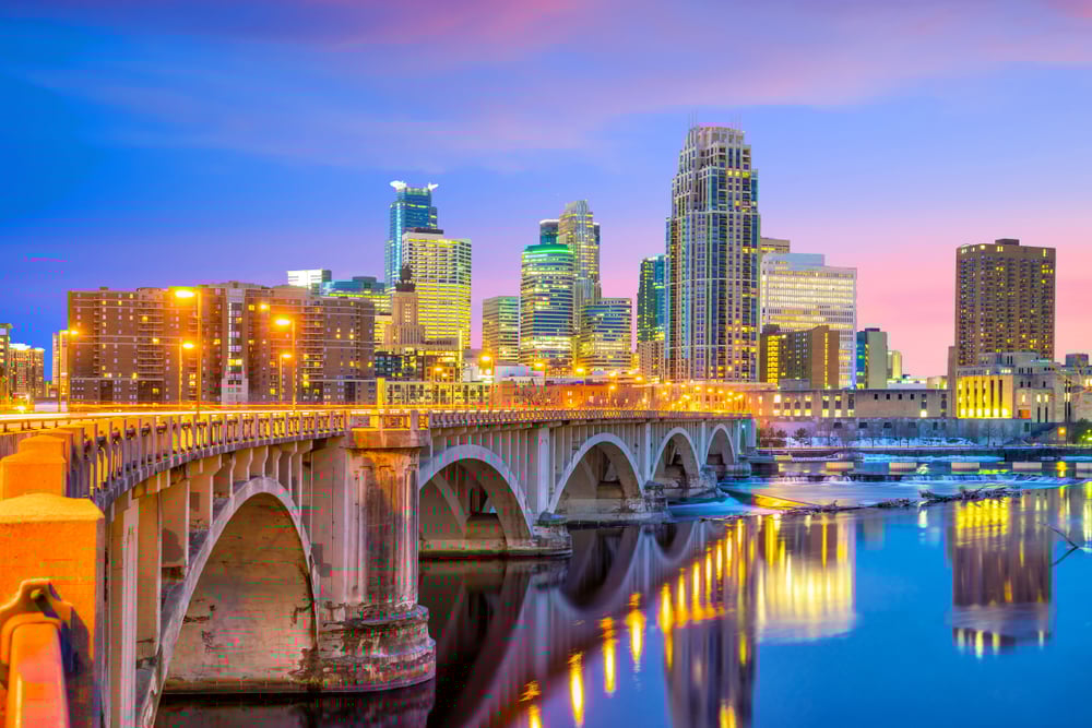 City skyline at dusk with illuminated bridge over calm river, colorful sky in background.