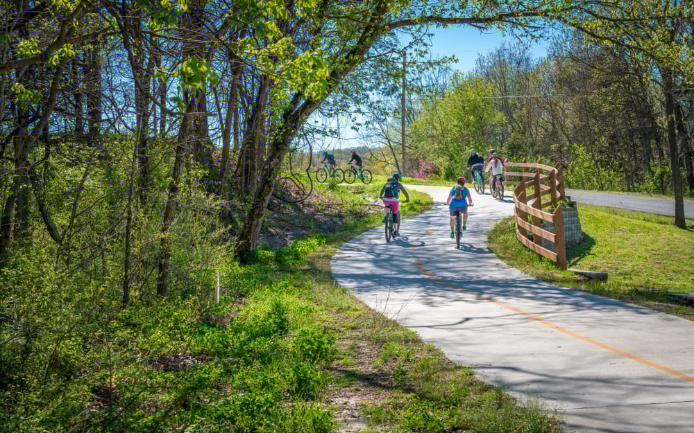 Winding park trail, cyclists, wooden fence framing curve, sunny midday light, editorial travel photo, with people.