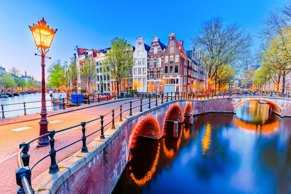 Illuminated canal, curved bridge, traditional buildings, and trees at twilight in a European city.