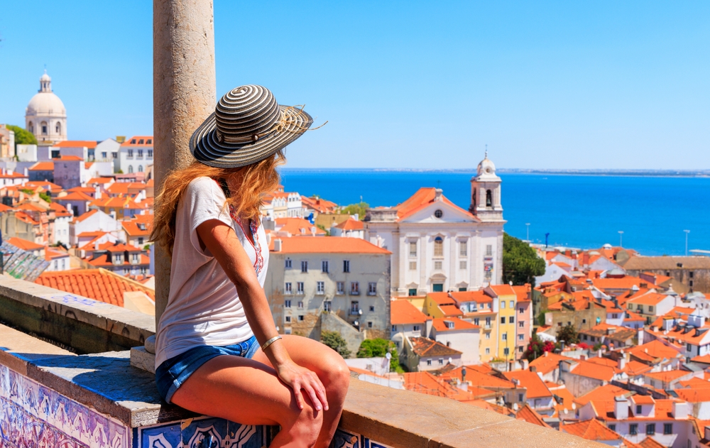 Woman in straw hat sits, overlooking Lisbon's orange rooftops and distant ocean under blue sky.