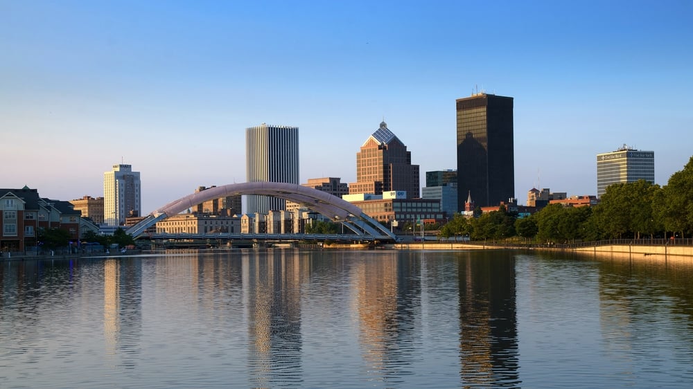 Skyline of Rochester, New York, with buildings, a large bridge, and reflective water in the foreground.
