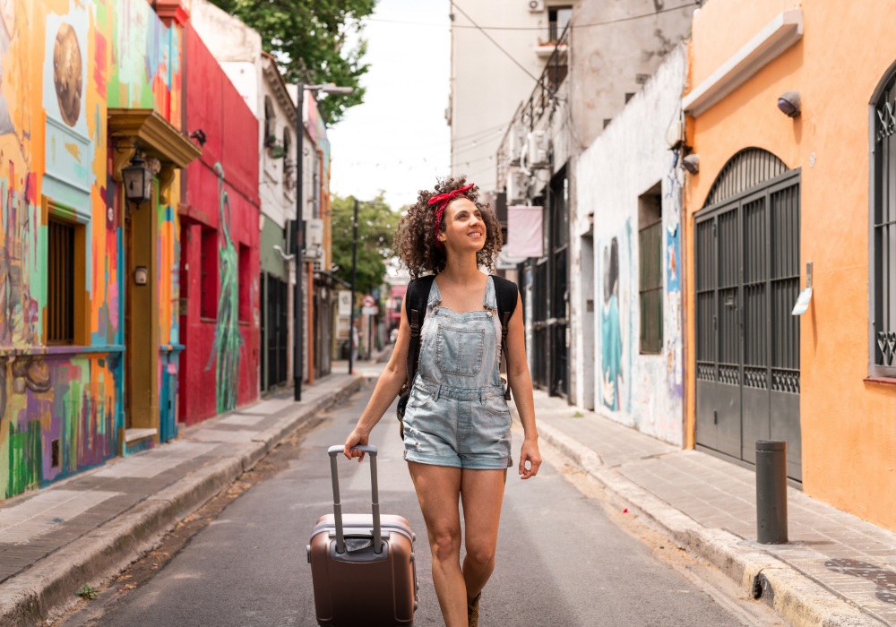 Mural-lined street, woman in denim overalls with flower crown, suitcase in hand, midday sun, editorial travel photo, single person.