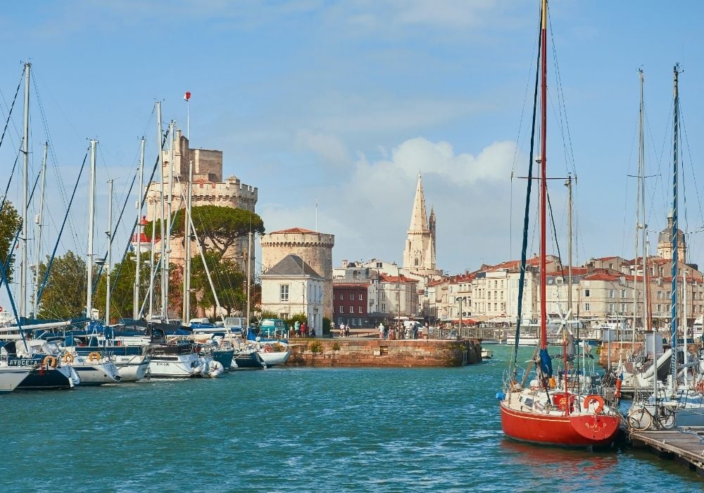 La Rochelle marina, sailboats moored by medieval towers, waterfront buildings, bright daylight, editorial travel photo, no people.
