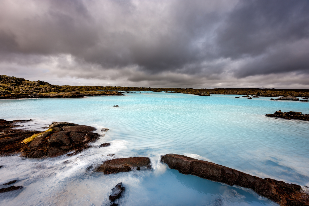 Cloudy sky above a turquoise geothermal lagoon surrounded by dark volcanic rocks.