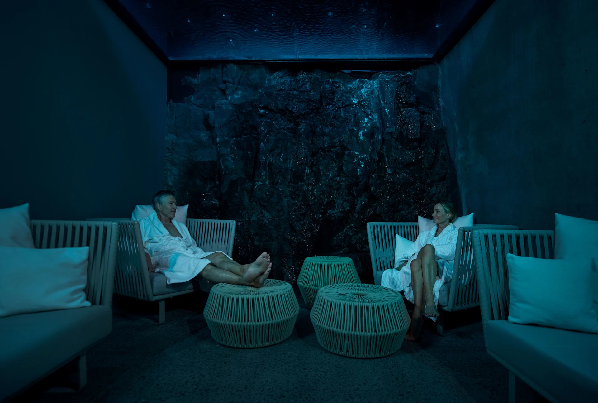 Two people in white robes sitting on chairs in a dimly lit room with rock wall.