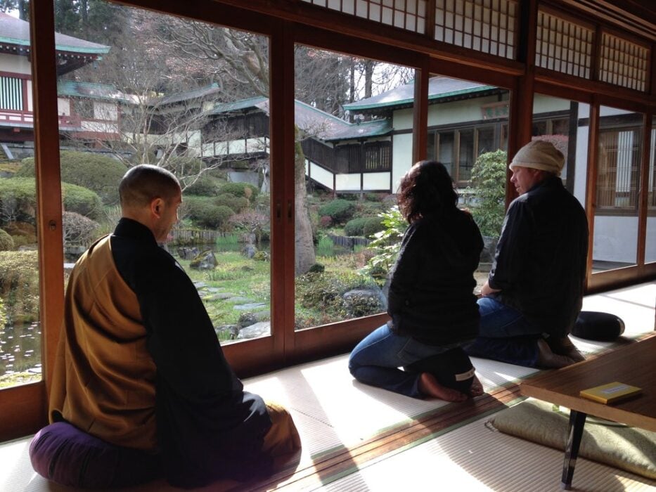 Three people kneeling indoors facing a garden through sliding glass doors.