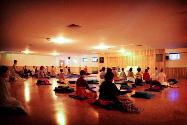 People sitting on meditation cushions in a dimly lit room with wood-paneled walls and ceiling lights.