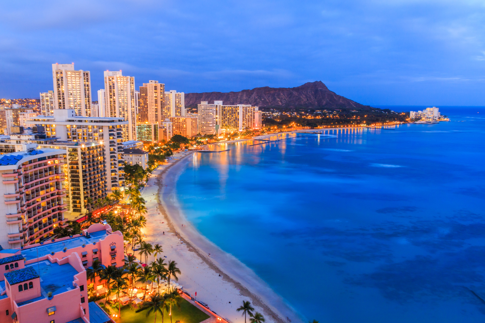 Waikiki Beach skyline at dusk with illuminated high-rise buildings and Diamond Head in the background.