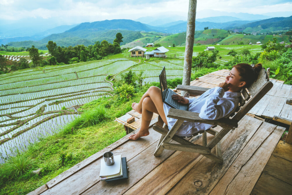 Woman sitting on wooden terrace with laptop, overlooking terraced rice fields and mountains.