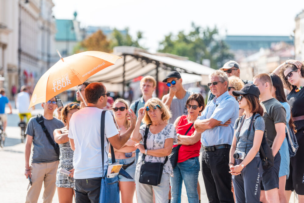 Tour guide holding orange umbrella speaks to a group of tourists on a city street.