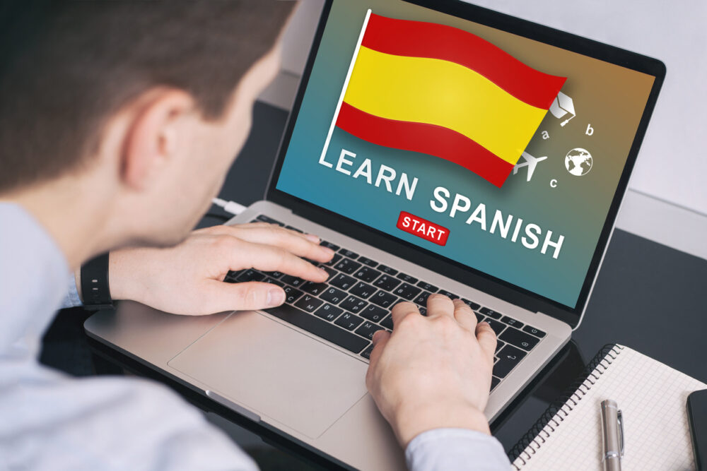 Person using a laptop showing "Learn Spanish" screen with a Spanish flag and start button.