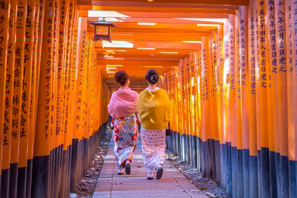 Two people in kimonos walk through orange torii gates with Japanese inscriptions.