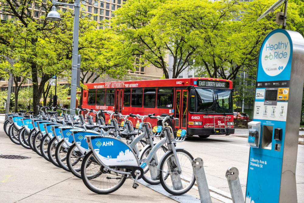 Rows of rental bikes and a red bus in front of green trees and buildings.