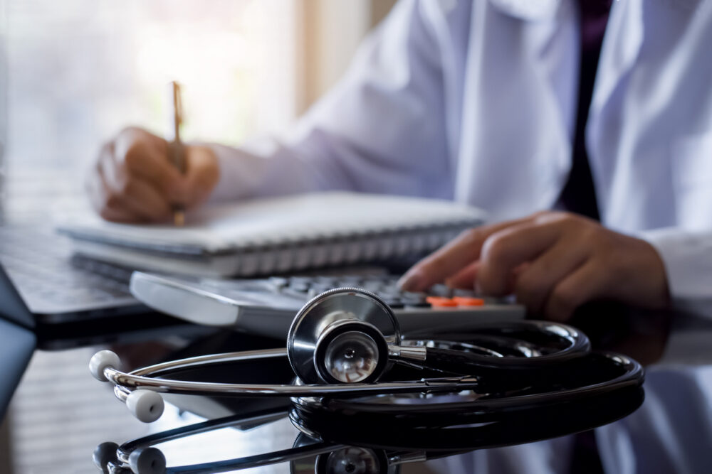 Stethoscope on a desk with person writing and using calculator in the background.