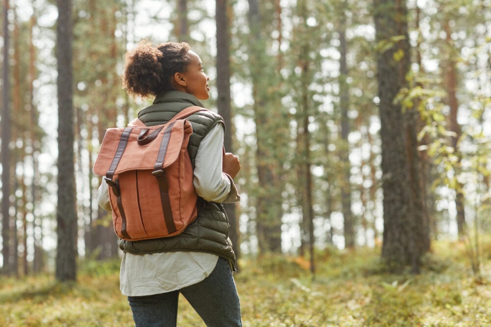 Person with orange backpack walks through a sunlit forest, wearing a vest and jeans.